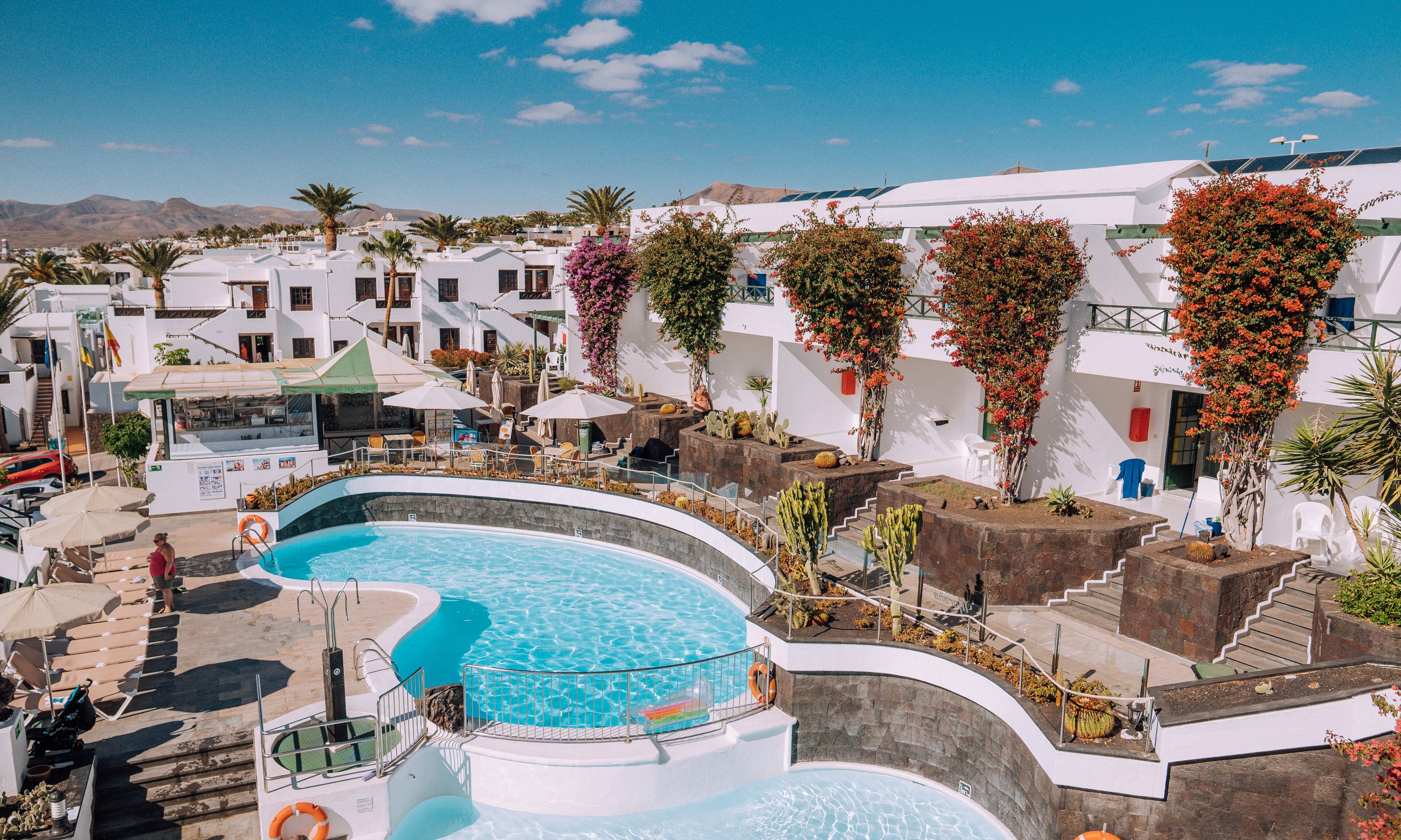 Un espace piscine au design incurvé, entouré de bâtiments blancs dotés de balcons et de cactus.