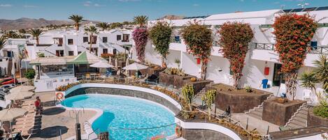 Un espace piscine au design incurvé, entouré de bâtiments blancs dotés de balcons et de cactus.