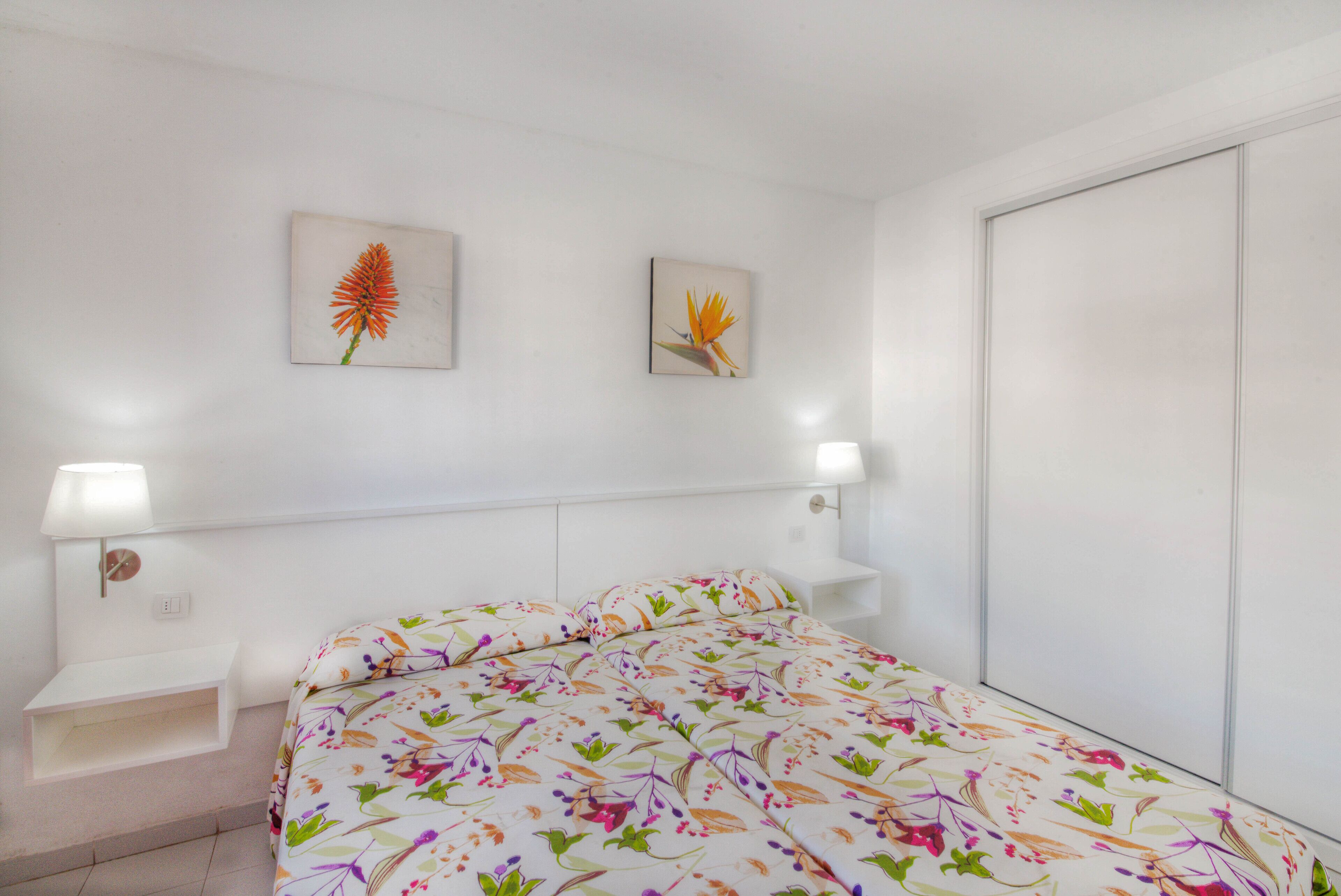 Un dormitorio con colcha estampada, dos cuadros en la pared, una mesita de noche con lámpara y un armario.