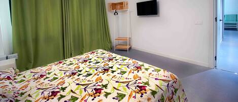 A room with a bed, green curtains, a mounted TV, and a small wooden shelf.