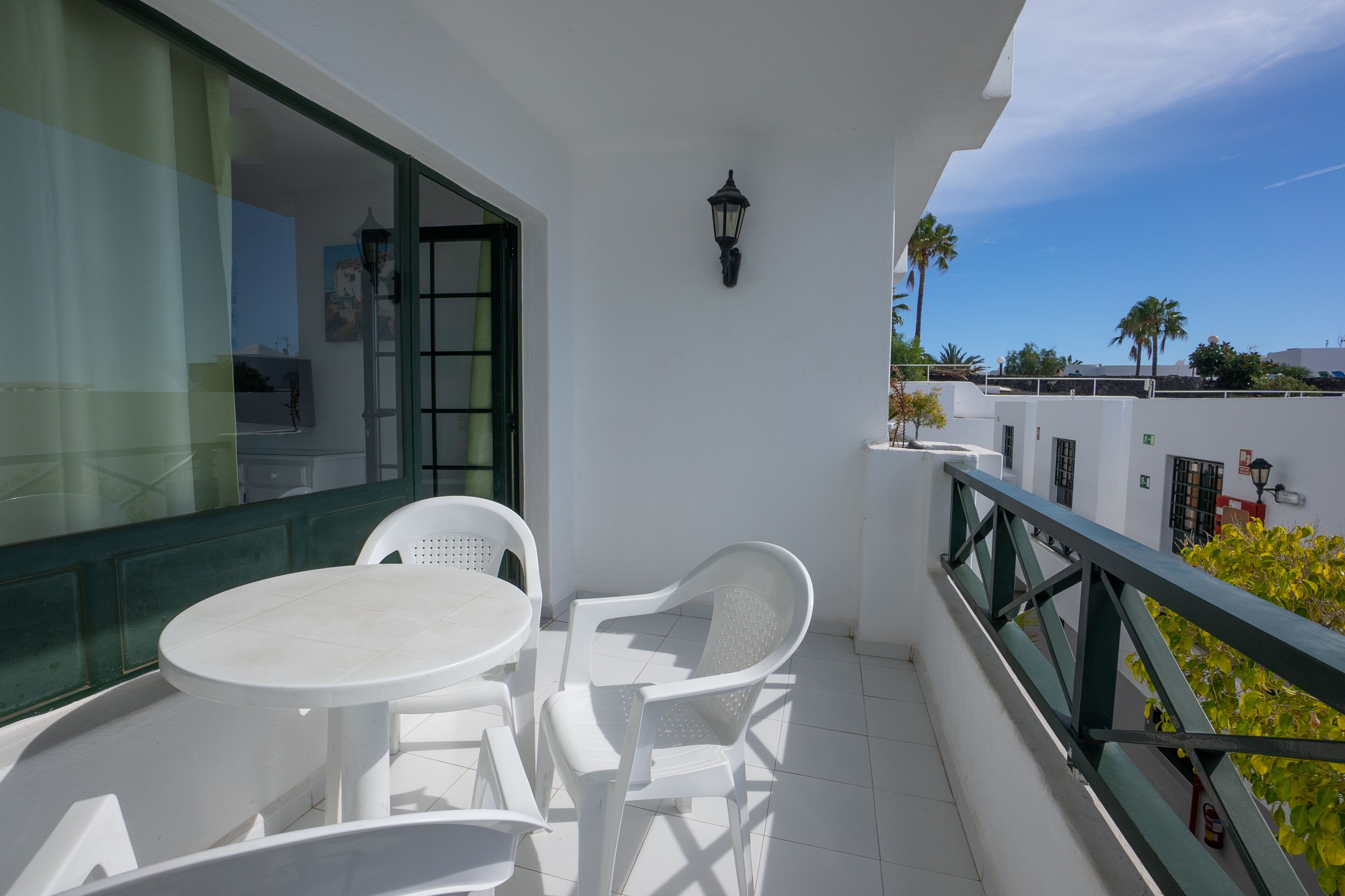 A balcony with a white table and chairs, a green railing, and a view of palm trees and buildings.
