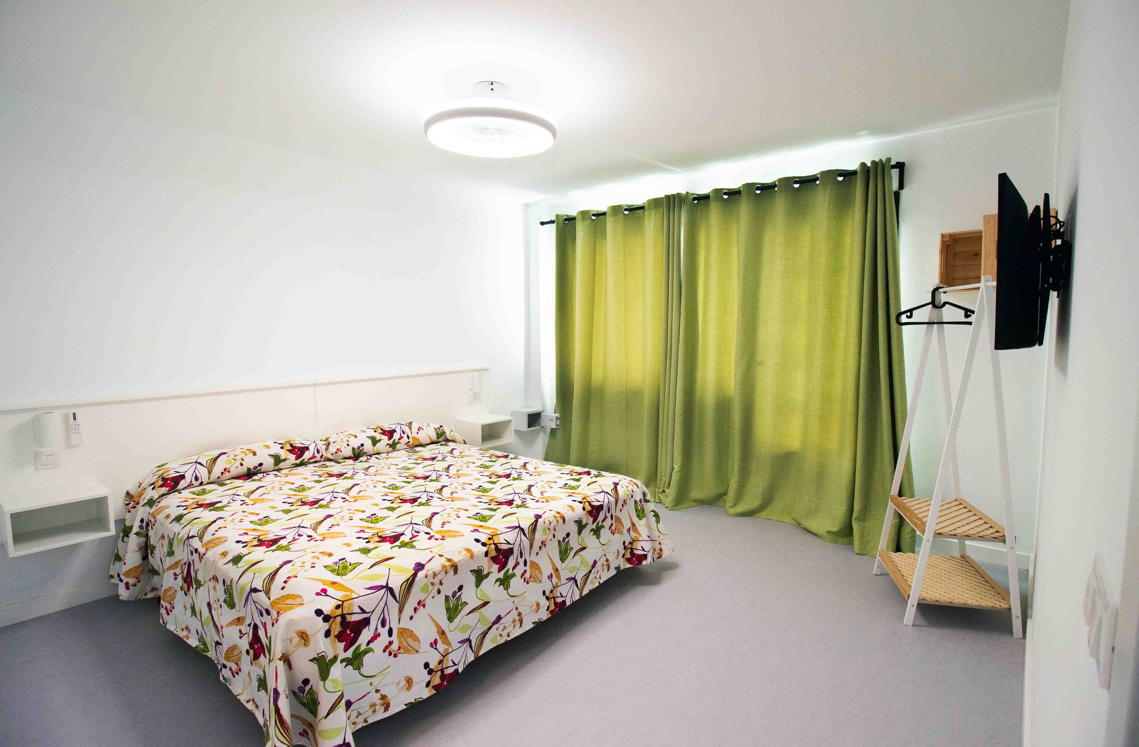 Una habitación con cama, un estante pequeño, perchero y ventana con cortinas verdes.