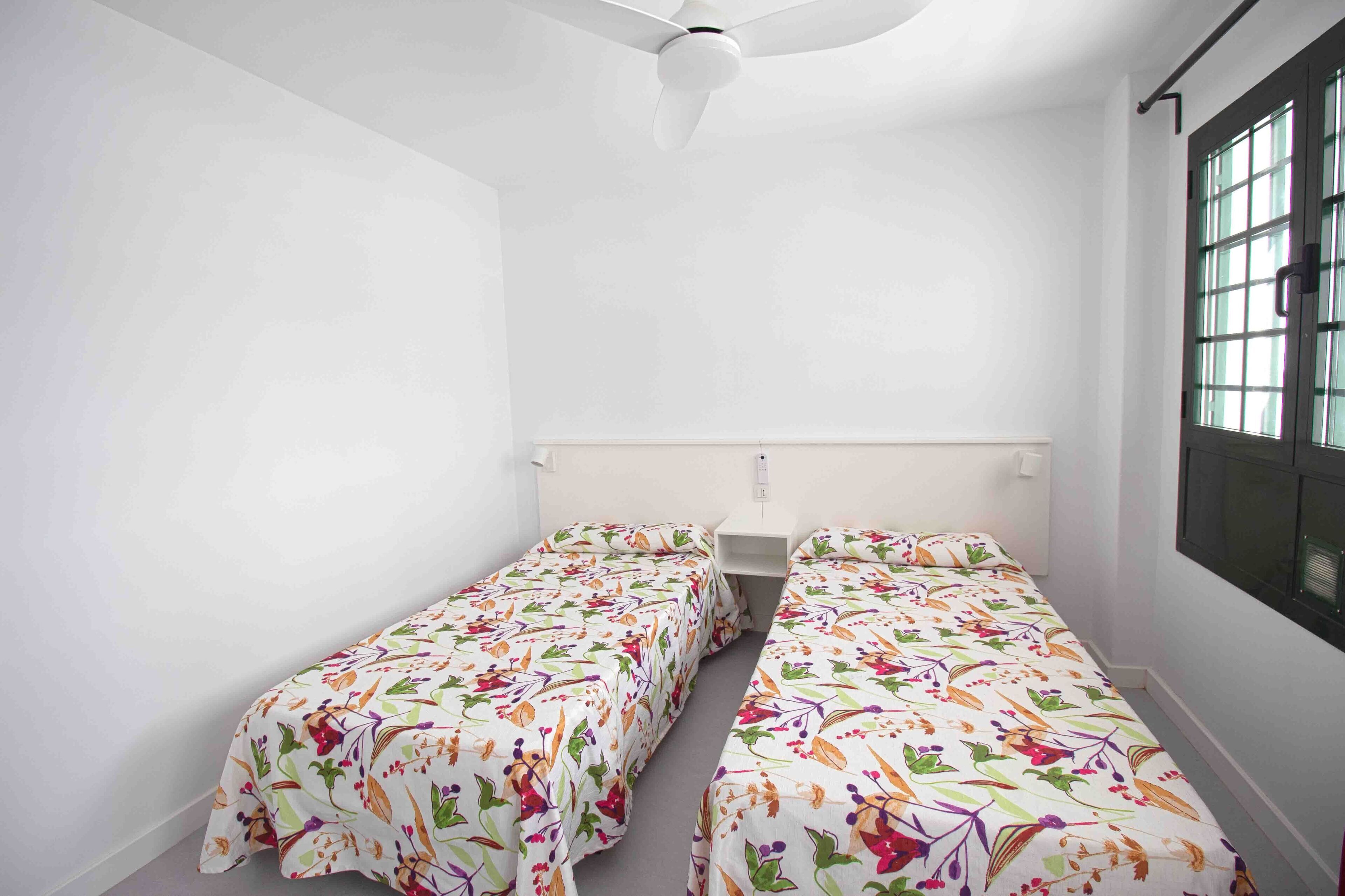 A small, single-room accommodation with two single beds, a ceiling fan, a window with blinds, and a white wall.