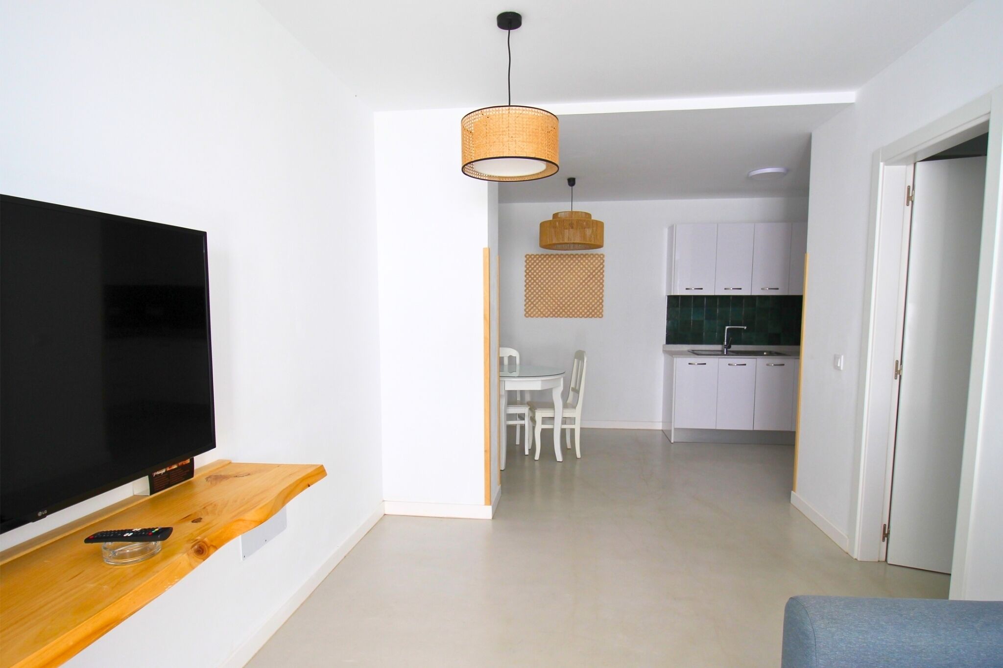Una sala moderna con un televisor de pantalla plana sobre una estantería de madera, un área de comedor con mesa y sillas, y una cocina con armarios blancos y salpicadero verde.