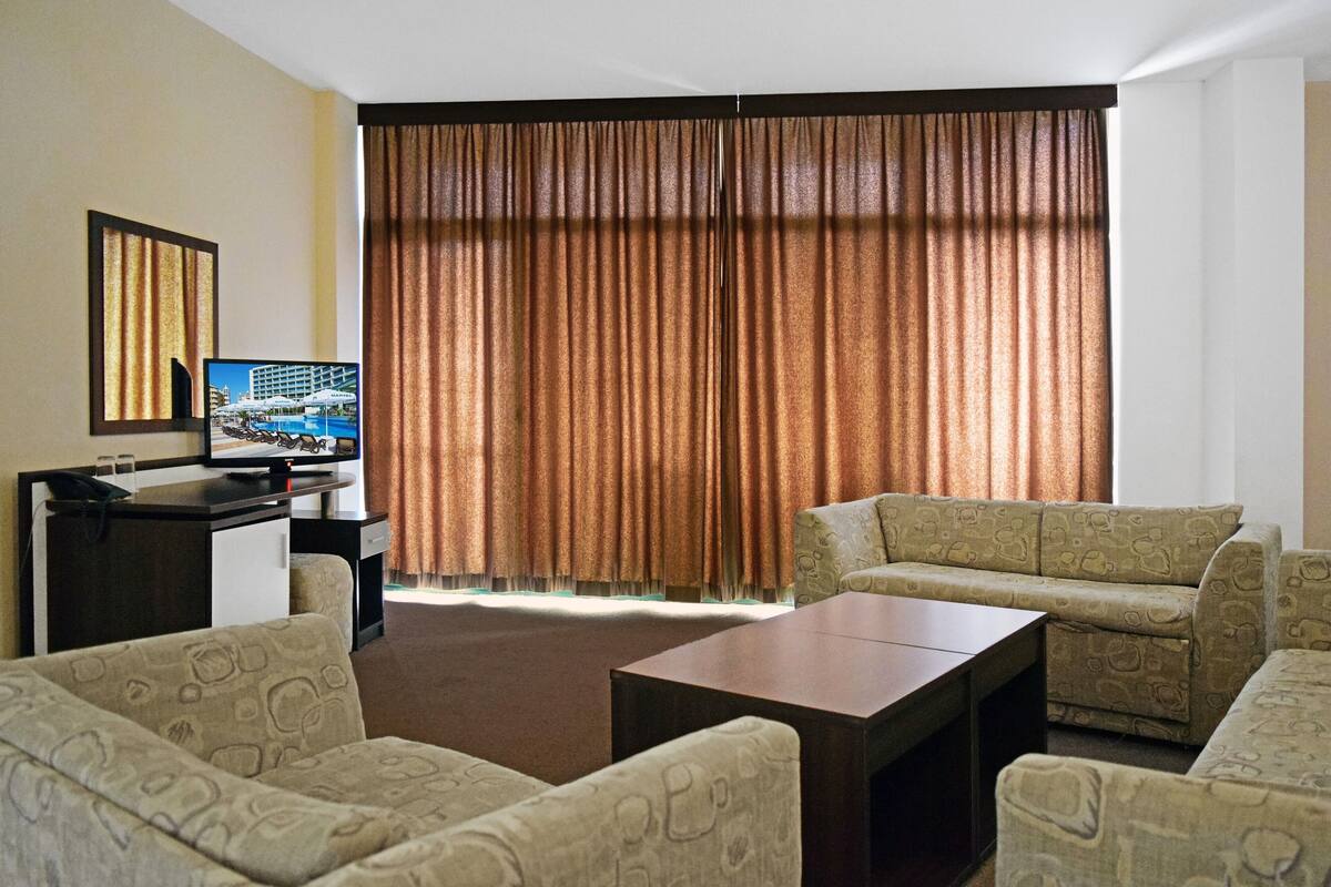 A hotel room with a large window, a desk with a computer, a sofa, and a coffee table.
