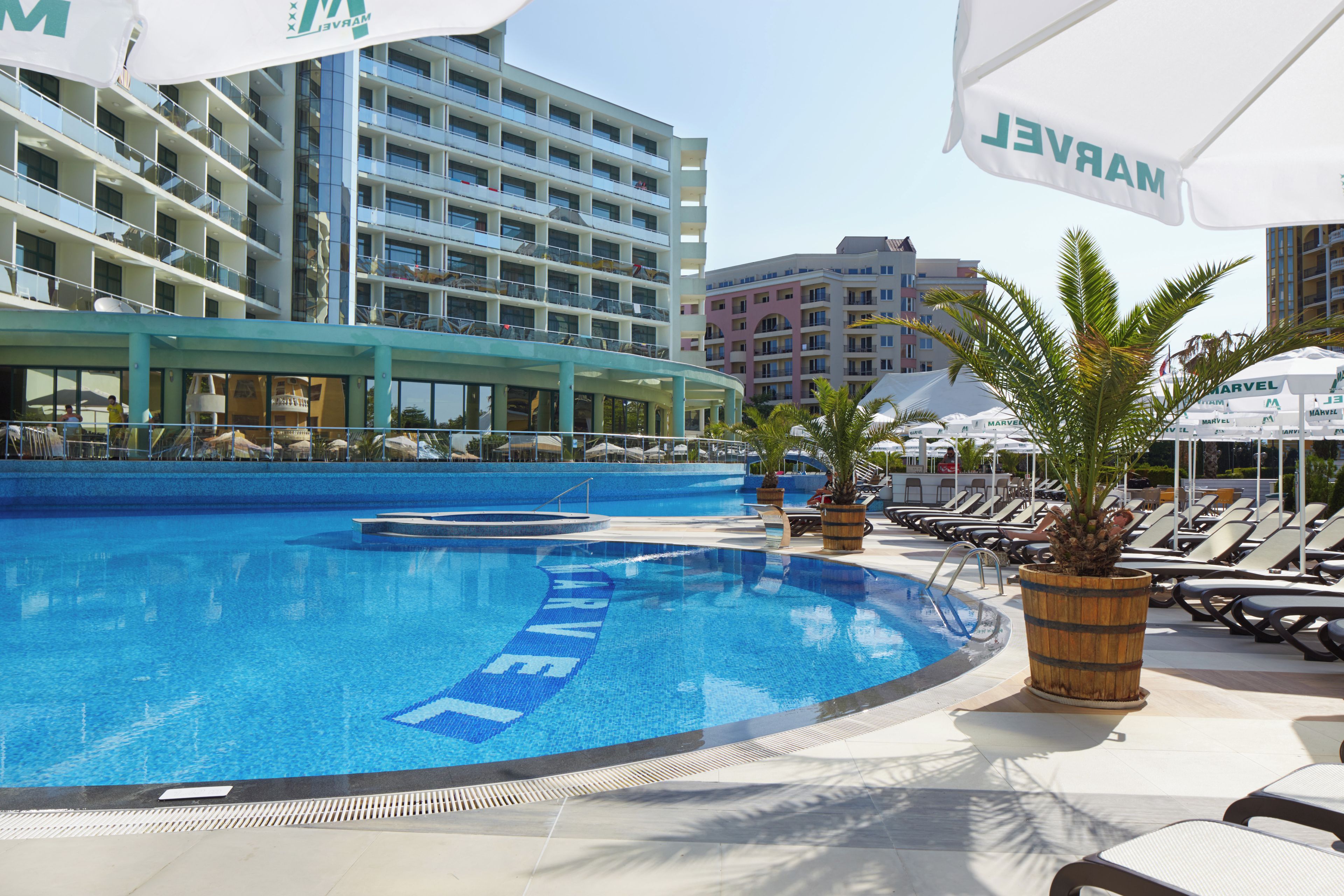 Zona de piscina de un hotel con sillones, sombrillas y un edificio con varios balcones al fondo.