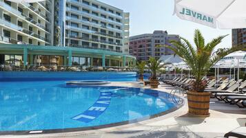 Zona de piscina de un hotel con sillones, sombrillas y un edificio con varios balcones al fondo.