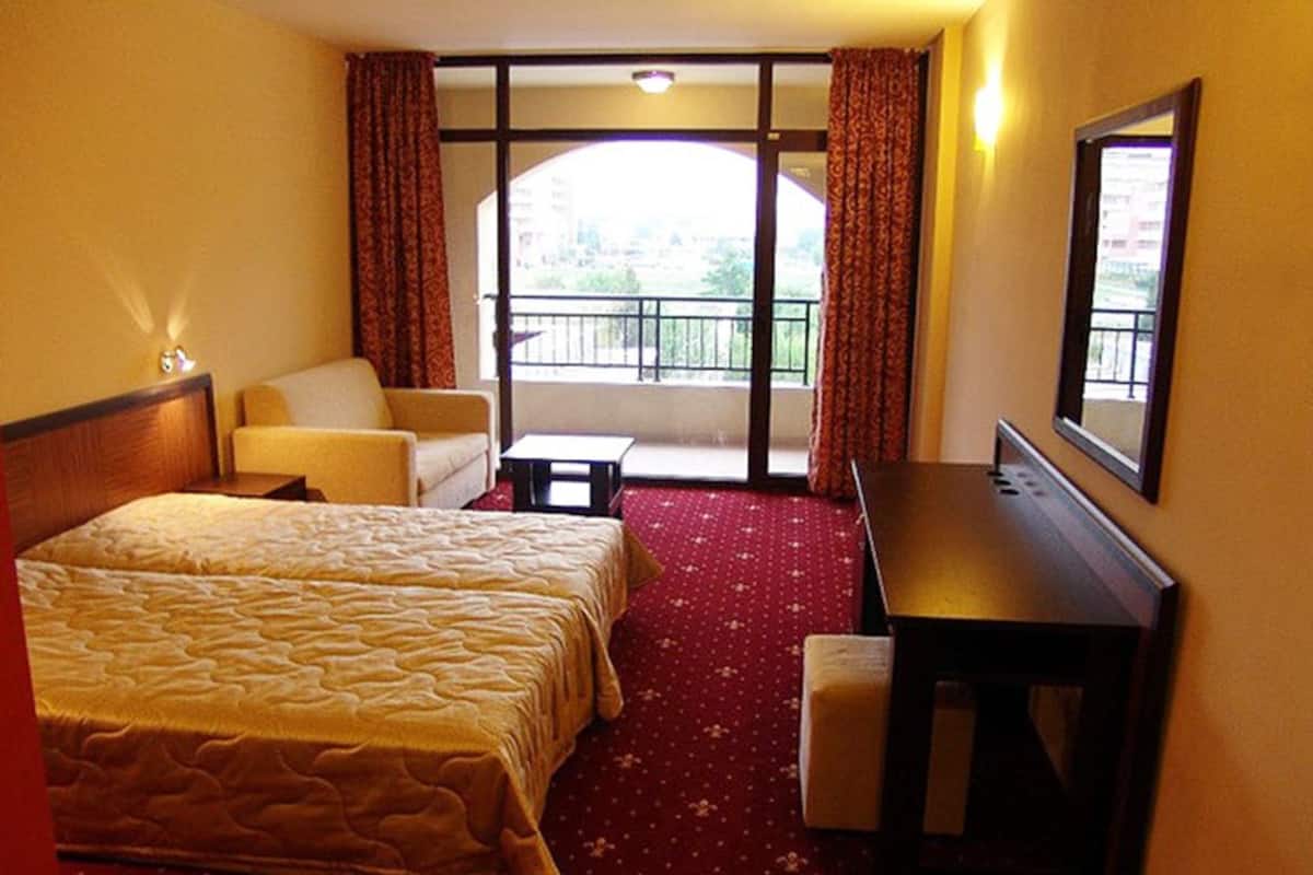 Quarto de hotel com cama, escrivaninha, cadeira e uma pequena mesa. Tem uma varanda com vista para a cidade.