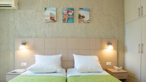 A hotel room with two beds, green bedding, white pillows, a wooden headboard, wall-mounted artwork, bedside lamps, a nightstand, and a wardrobe. - Alkionis (Corfu)