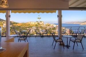 Terraza al aire libre con mesas y sillas, con vistas a un pueblo costero y al mar.