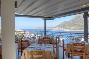 An outdoor dining area with tables set for two, overlooking a coastal landscape with mountains and the sea. - Aparthotel Athina (Mylopotamos)