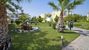 A well-maintained garden with palm trees, a circular stone feature with rocks and a teddy bear, and a paved walkway. - Corali Hotel (Kos)