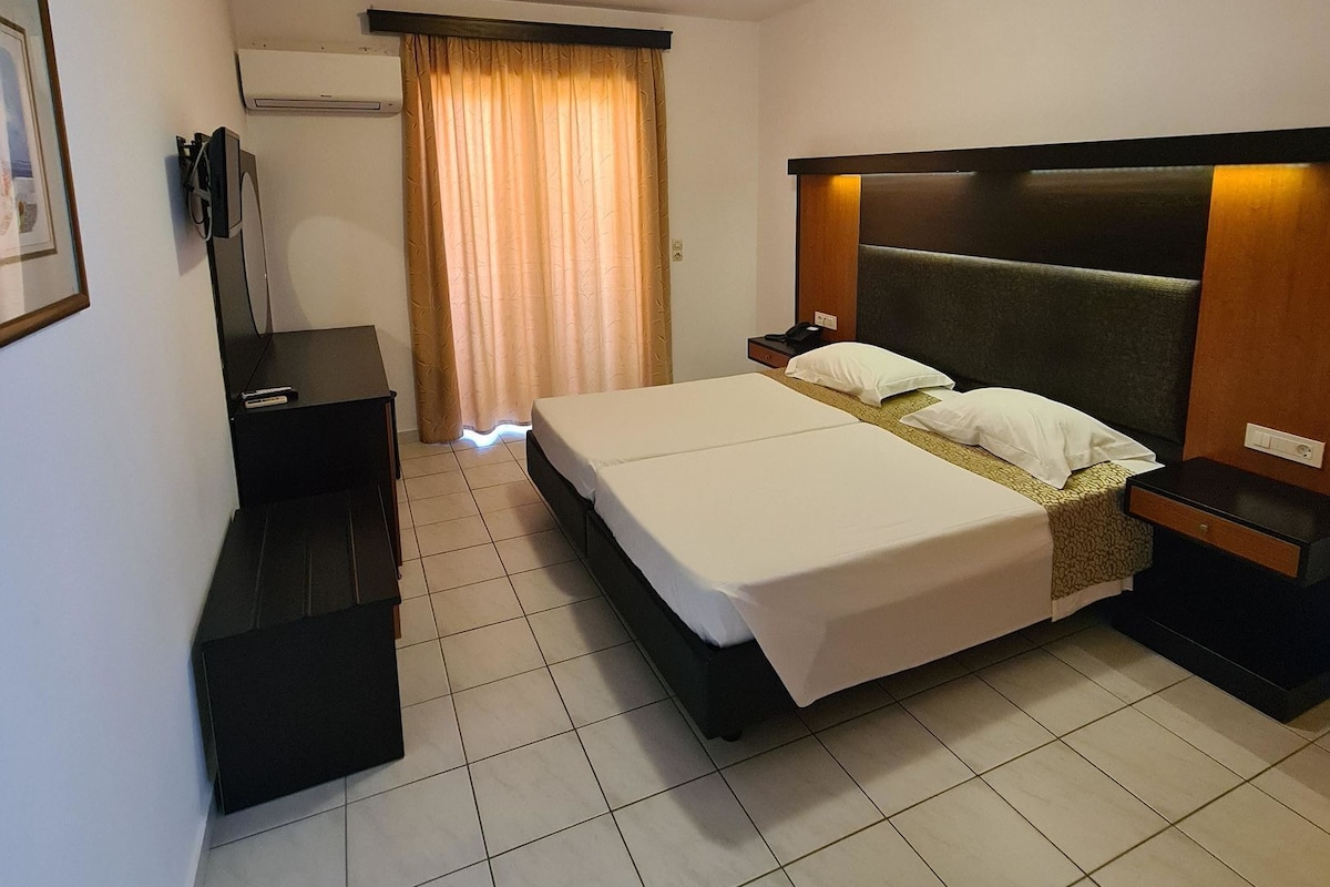 Une chambre d’hôtel comprenant un lit, des tables de chevet, un bureau, une chaise, une armoire et une fenêtre avec des rideaux.