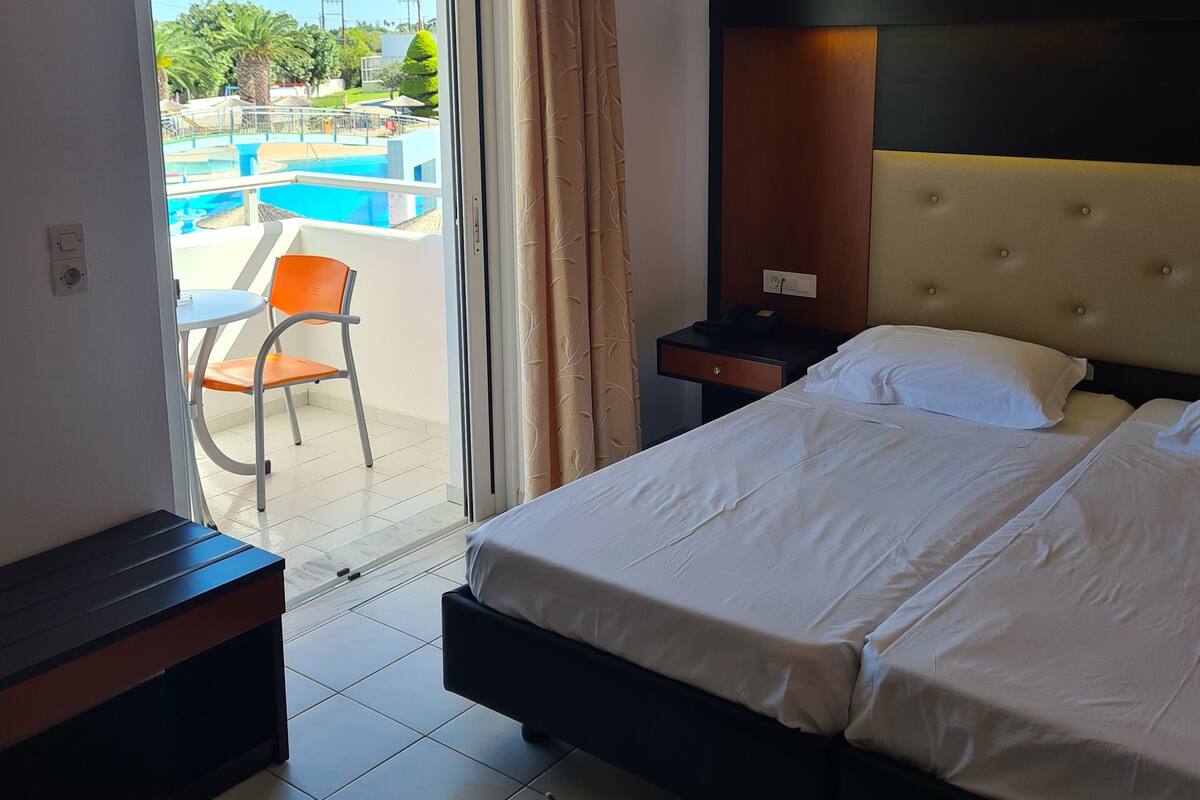 A hotel room with a bed, a bedside table, a nightstand, a window with curtains, and a view of a swimming pool and greenery outside.