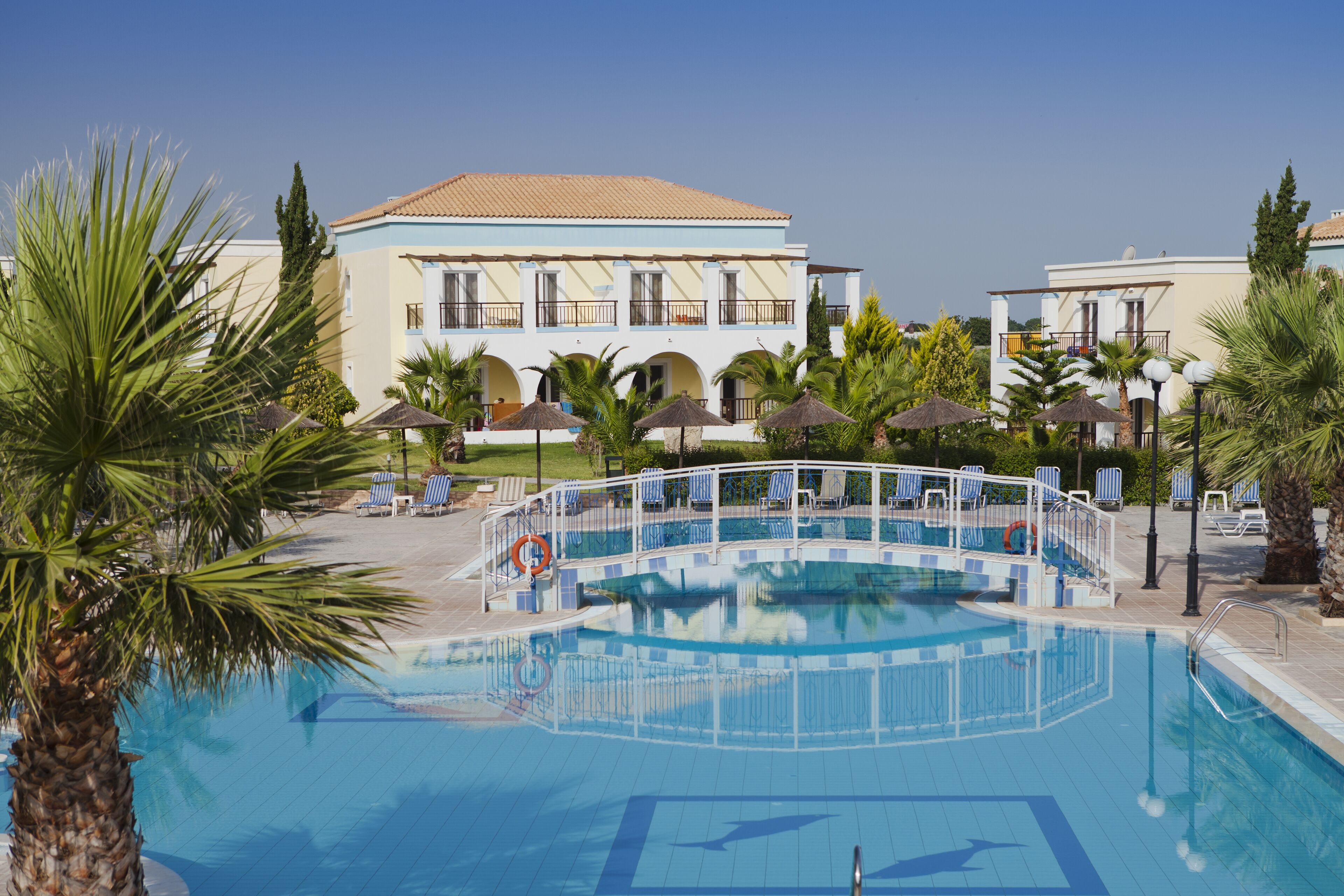 A pool area with lounge chairs and umbrellas, surrounded by palm trees and a building with a tiled roof.