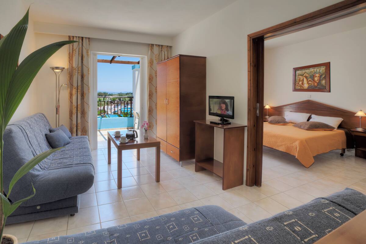A hotel room with a sofa, a small table with a cup and a book, a bed with an orange bedspread, a television, and a painting on the wall.