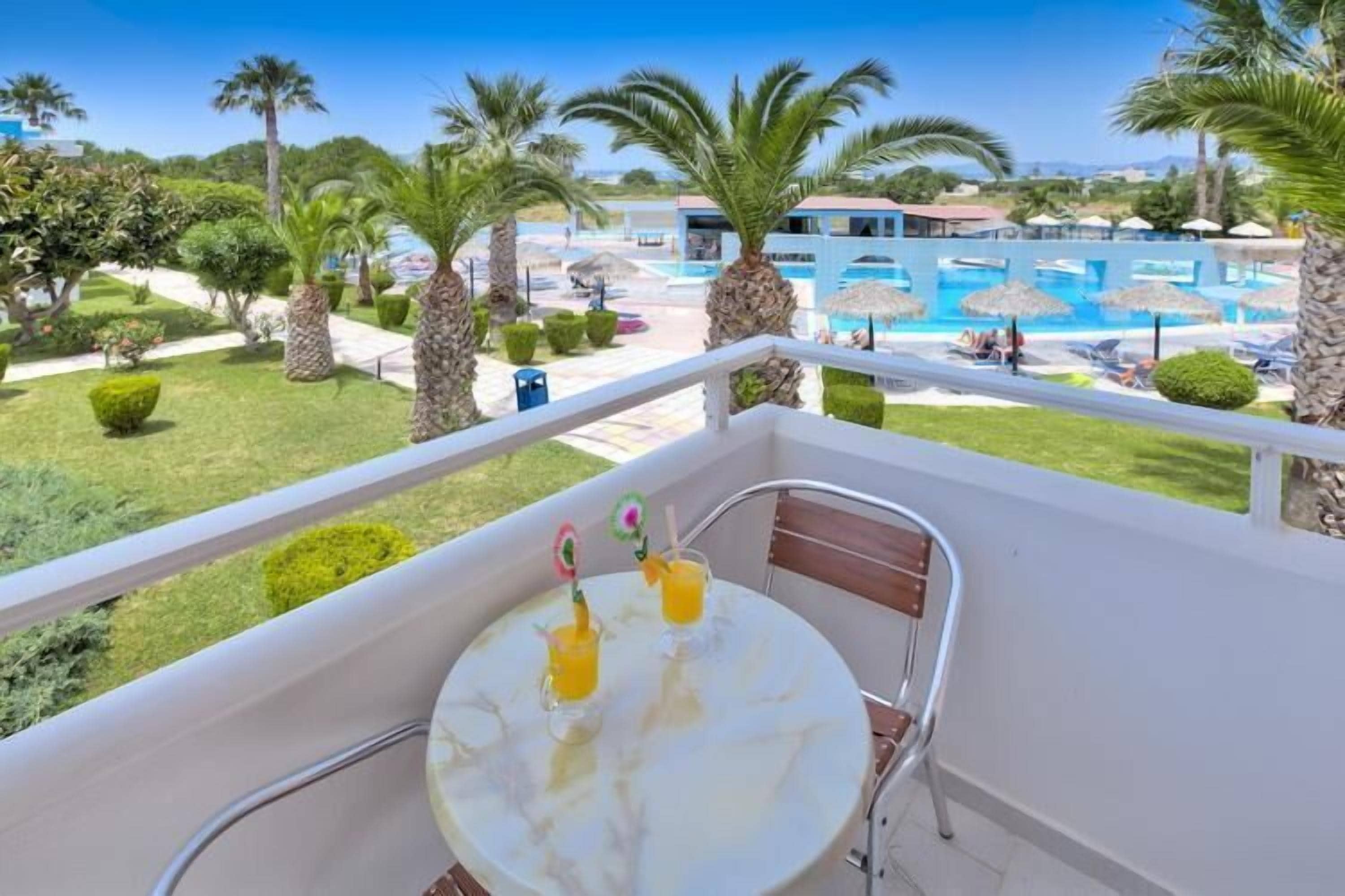 Un balcón con una mesa, dos bebidas y vistas a una piscina rodeada de palmeras y sombrillas.