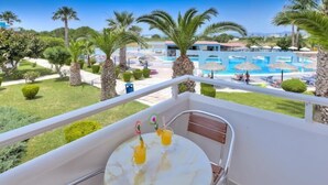 A balcony with a table, two drinks, and a view of a pool area with palm trees and umbrellas. - Corali Hotel (Kos)