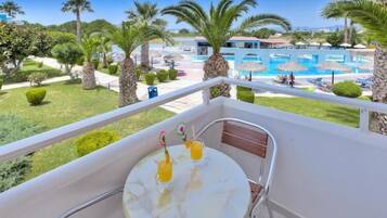 Un balcón con una mesa, dos bebidas y vistas a una piscina rodeada de palmeras y sombrillas.