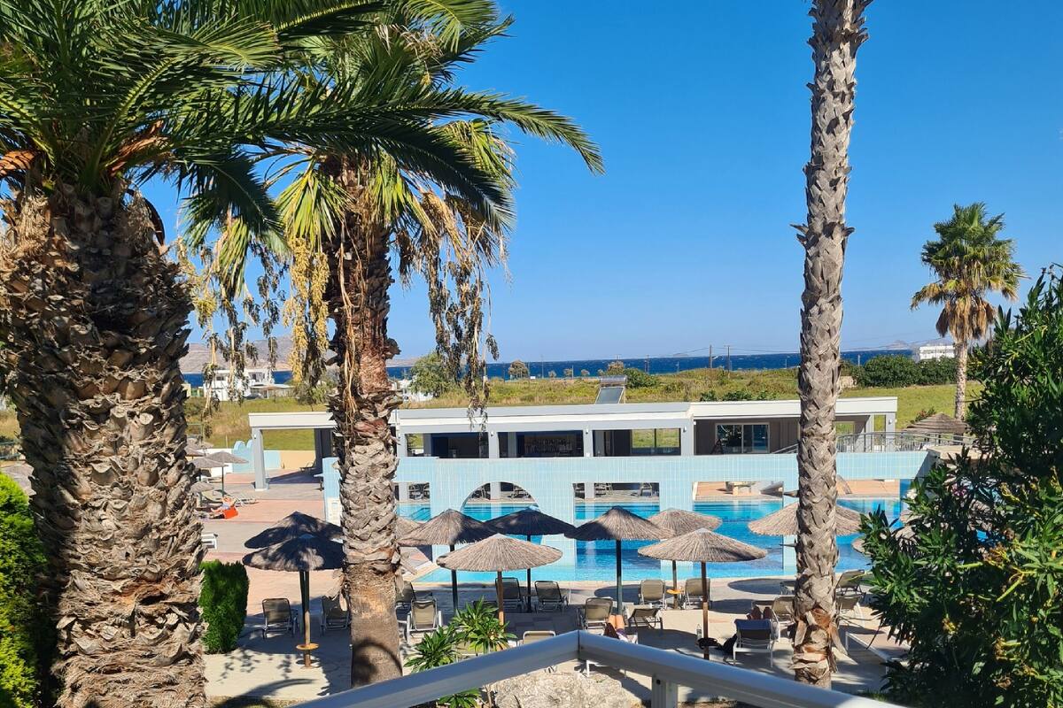 A balcony with a view of a pool area, palm trees, and a building with a blue facade.