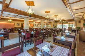 Un comedor espacioso con múltiples mesas y sillas, vigas de madera en el techo y candelabros colgantes.