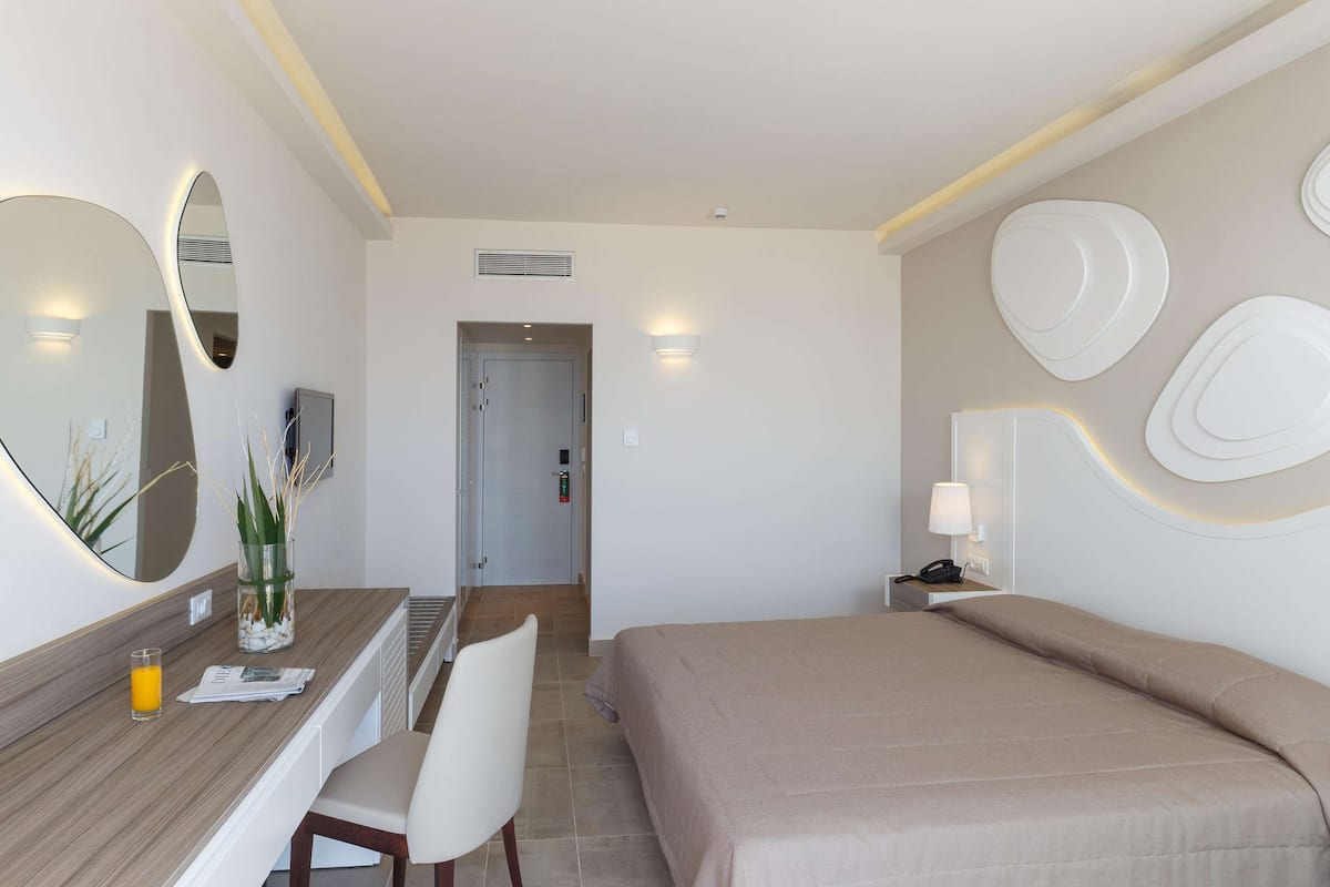A hotel room with a bed, a desk with a chair, a mirror, and a wall-mounted lamp.
