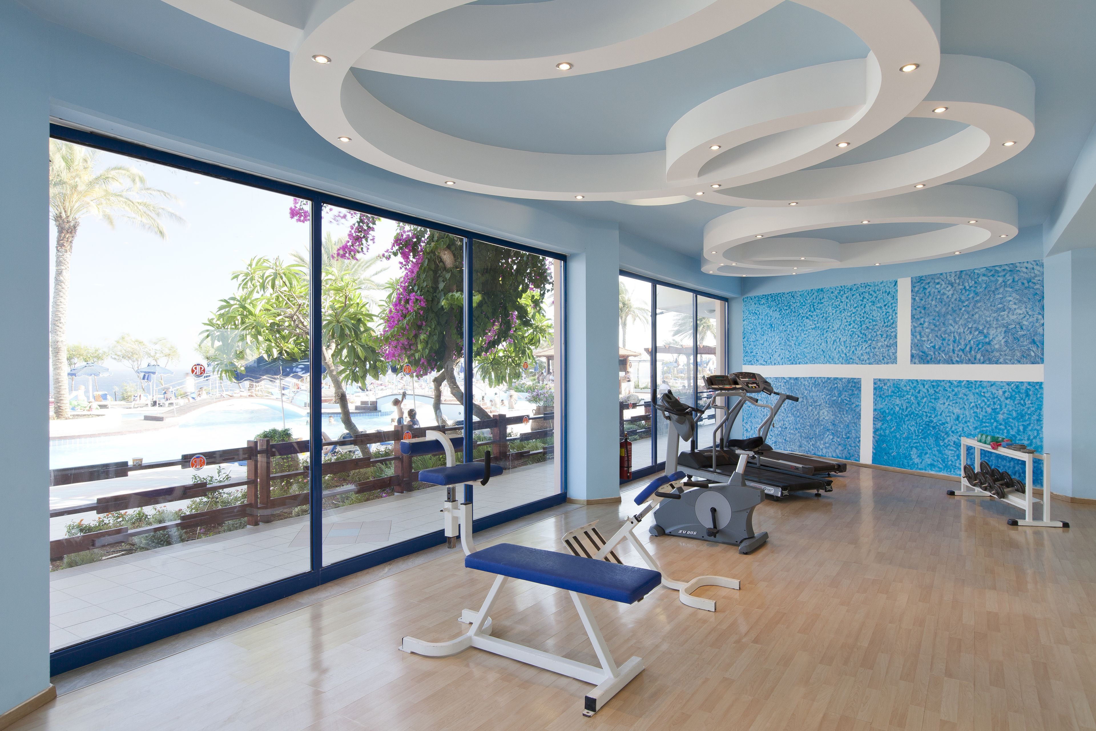A gym with exercise equipment, large windows, and a view of a pool and palm trees.