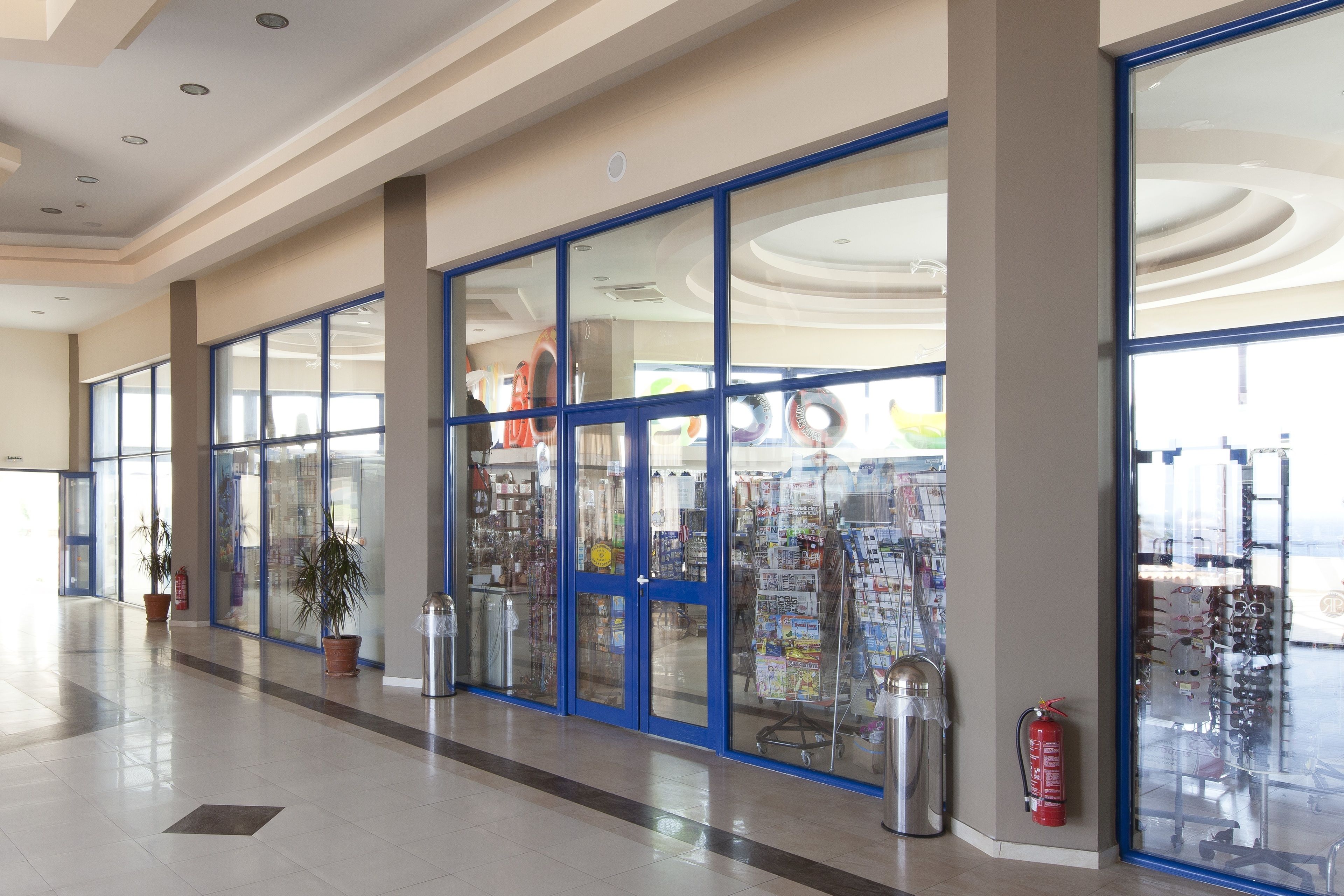 A spacious corridor with large glass doors, potted plants, and a fire extinguisher.