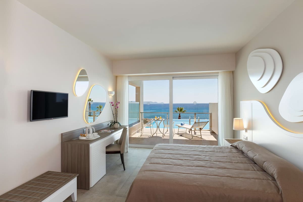 A modern hotel room with a bed, desk, and a large window overlooking the sea.