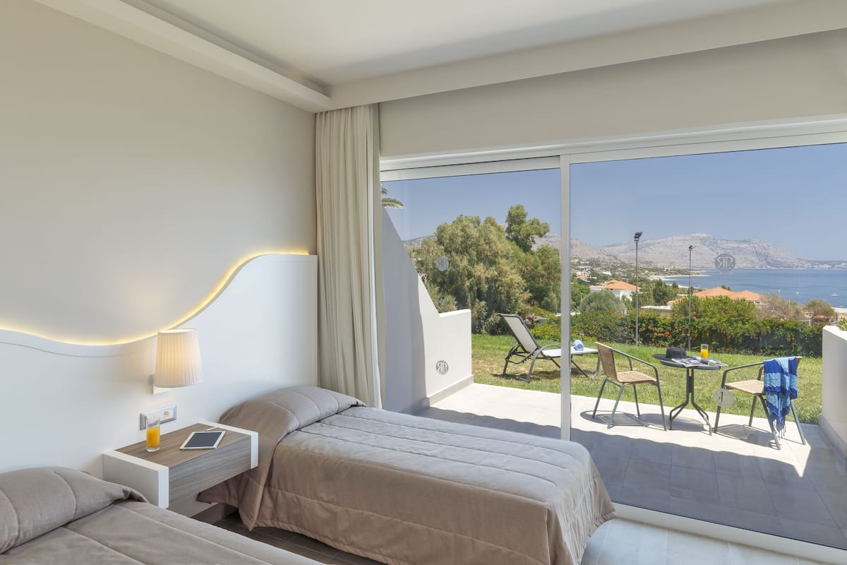 A hotel room with two beds, a bedside table with a lamp, a tablet, and a glass of orange juice. The room has a large sliding glass door leading to an outdoor area with a table, chairs, and a view of the sea and hills.