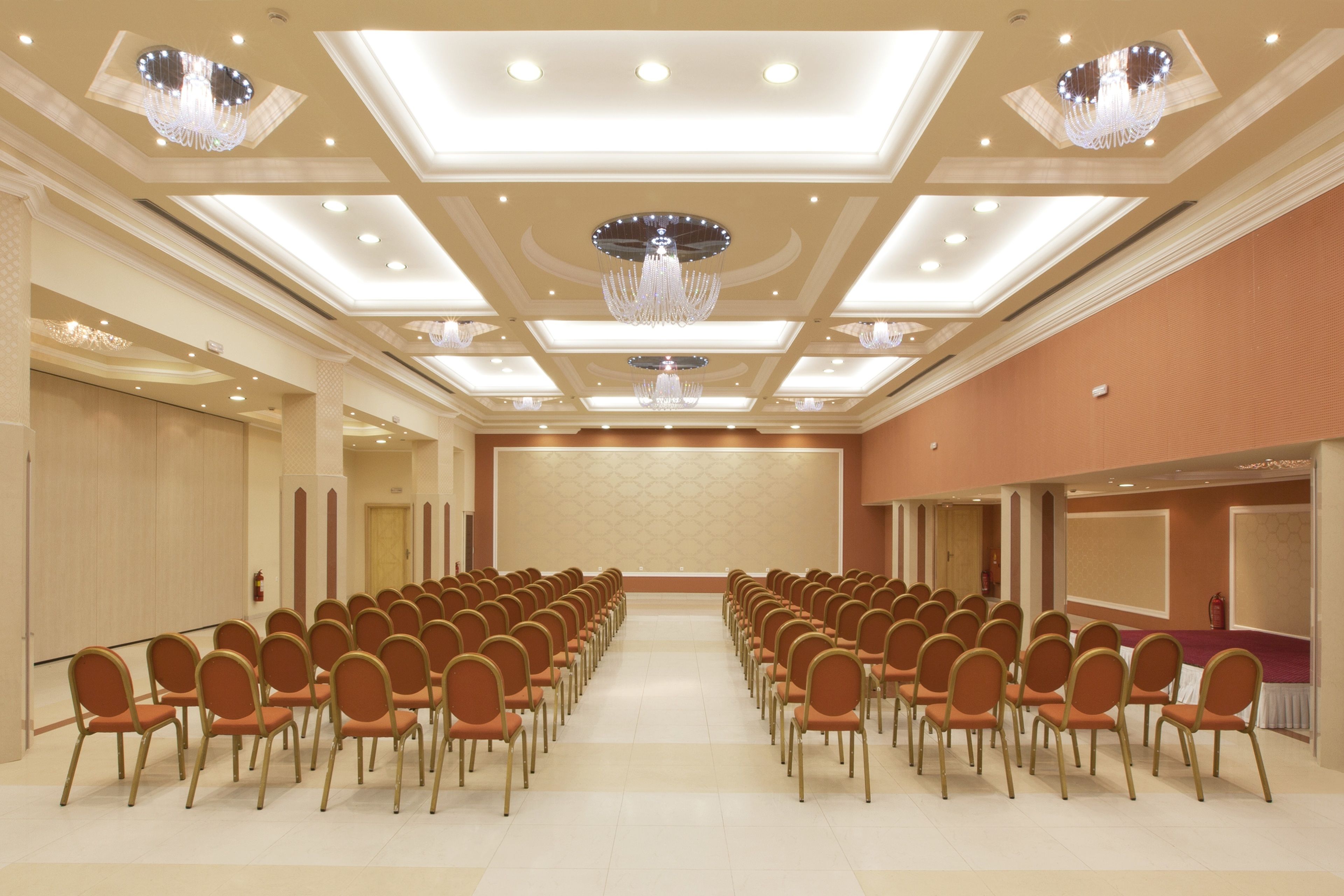 A conference room with rows of chairs facing a stage, recessed lighting, and chandeliers.
