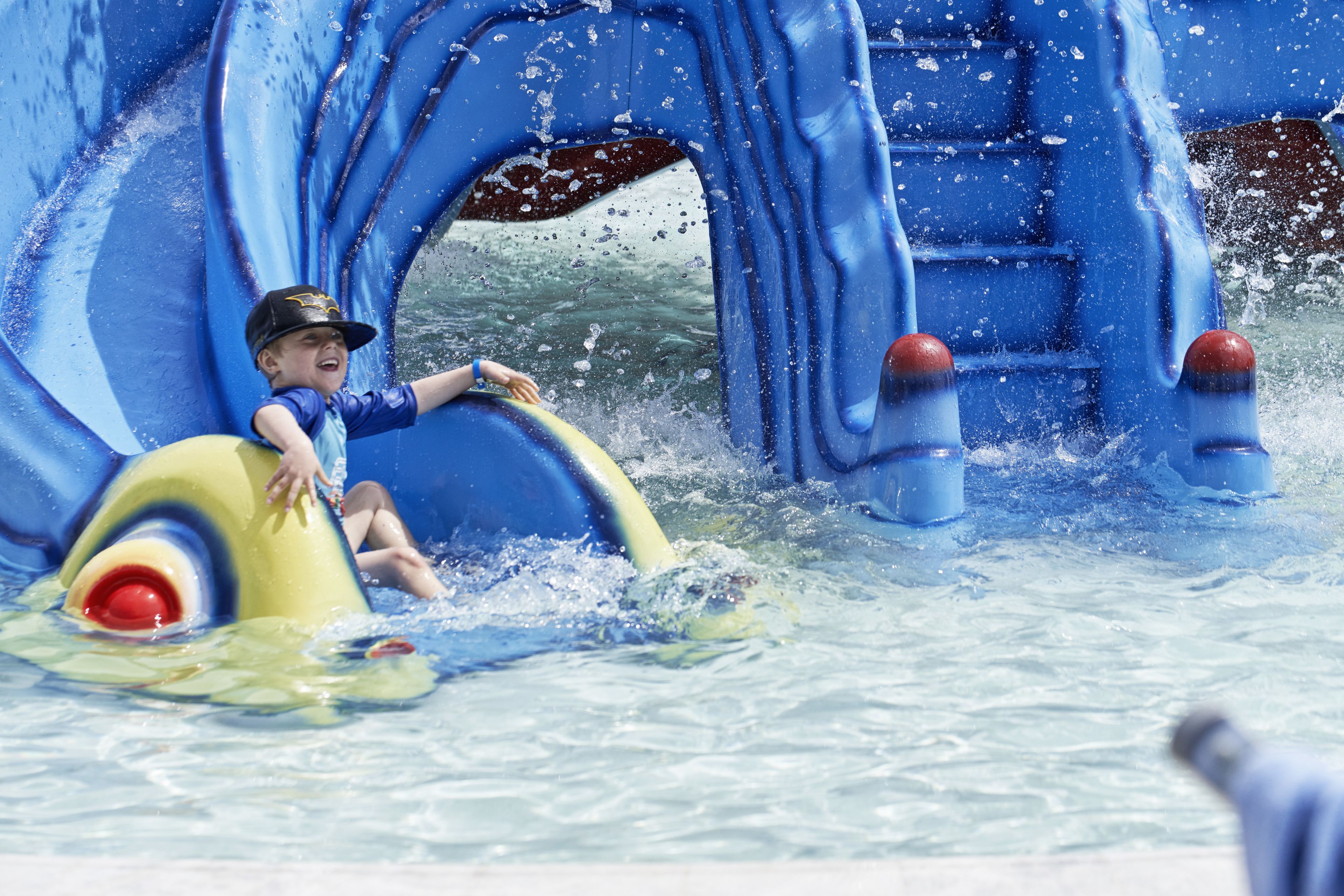 A child on a yellow raft in a water play area with blue slides and water features.