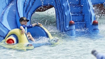 A child on a yellow raft in a water play area with blue slides and water features.