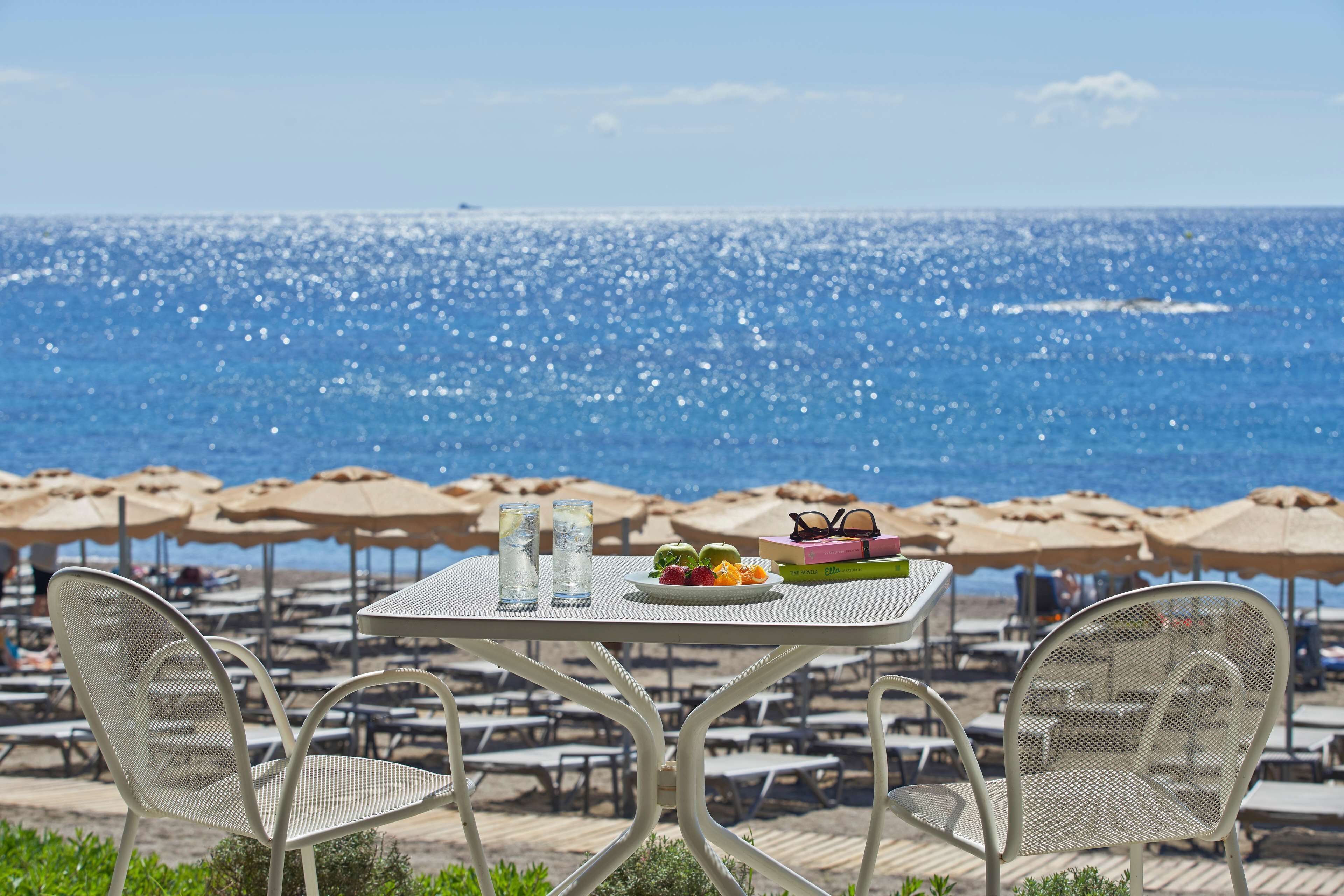 Ein Tisch mit einer Fruchtschale, Gläsern und einem Buch mit Blick auf einen Strand mit Sonnenschirmen und Liegestühlen.