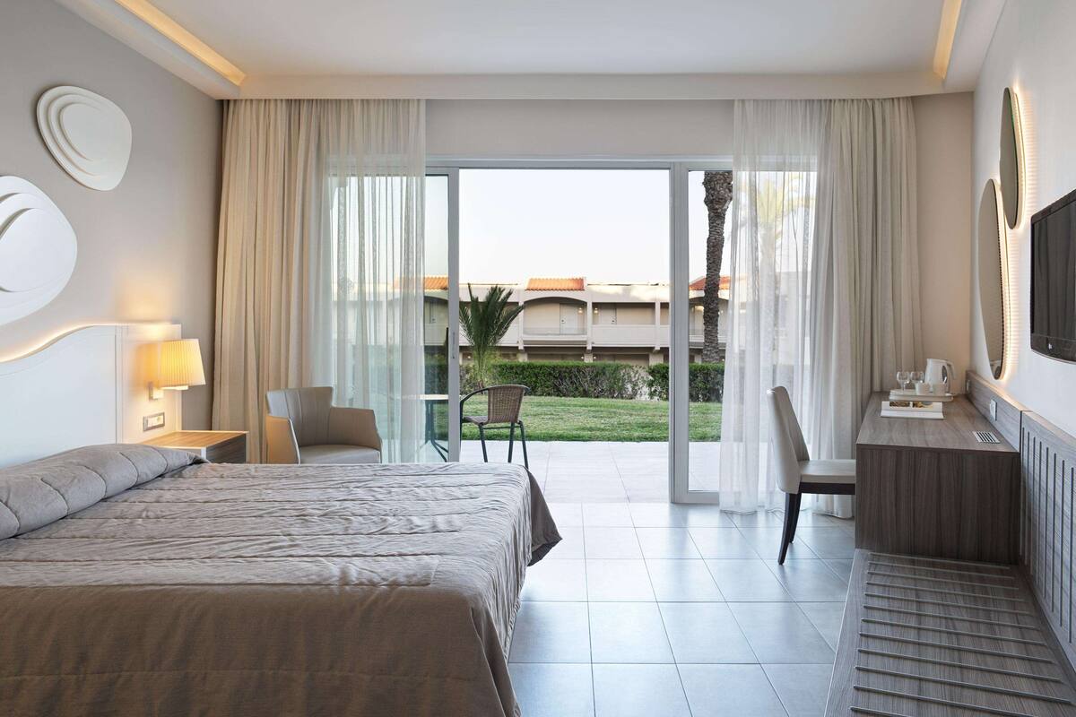 A modern hotel room with a large bed, a desk with a chair, a TV, and a view of a building and greenery outside.