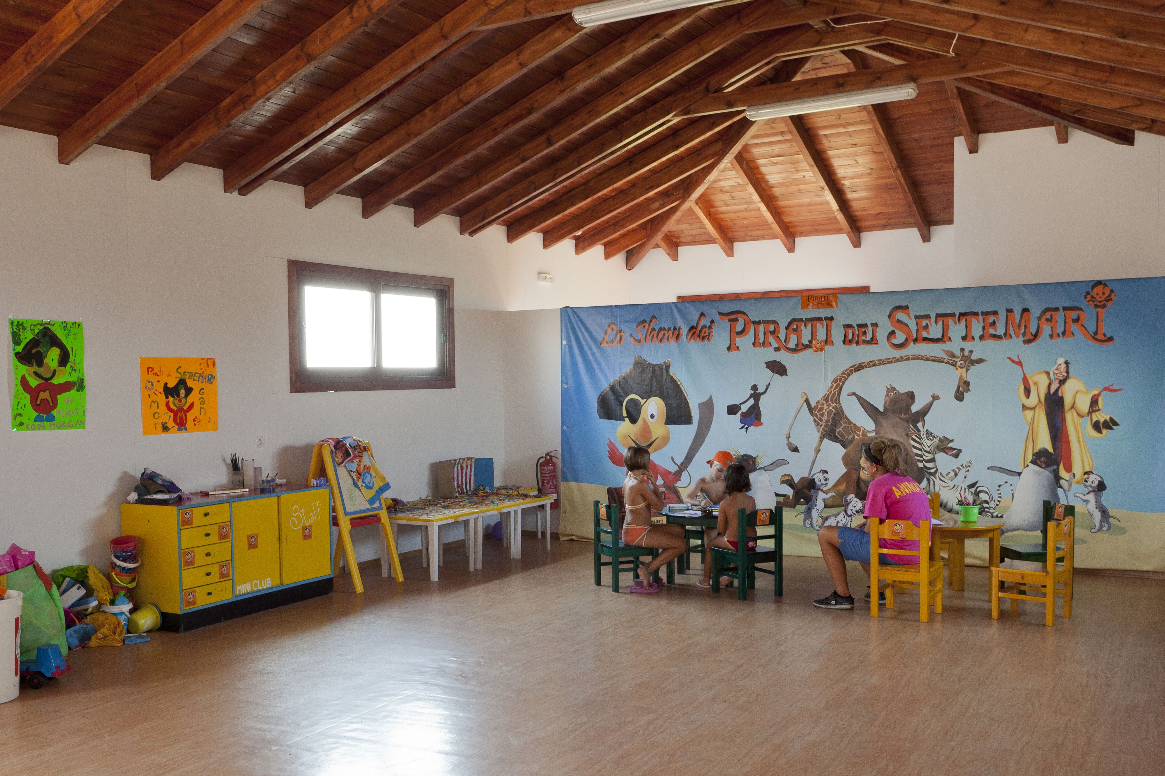 A room with wooden beams on the ceiling, a large mural on the wall, a yellow cabinet, and a table with chairs.