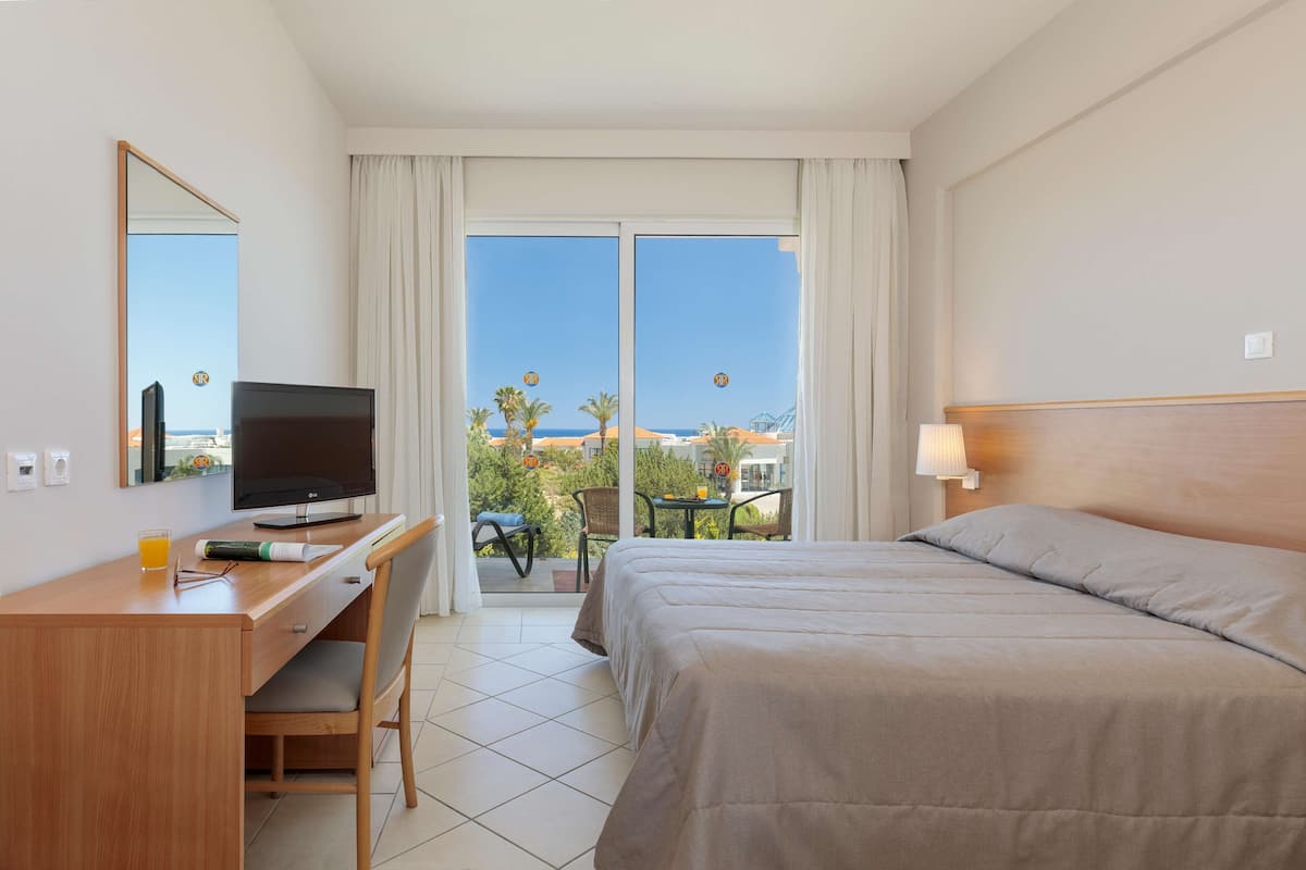 A hotel room with a bed, a desk with a TV, a chair, a table with a lamp, and a balcony with a view of palm trees and buildings.