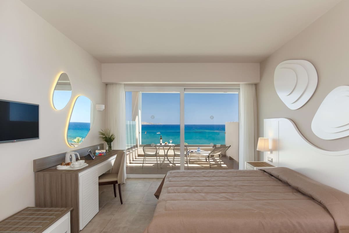 A hotel room with a bed, a desk with a TV, a balcony with a view of the sea, and a large mirror.