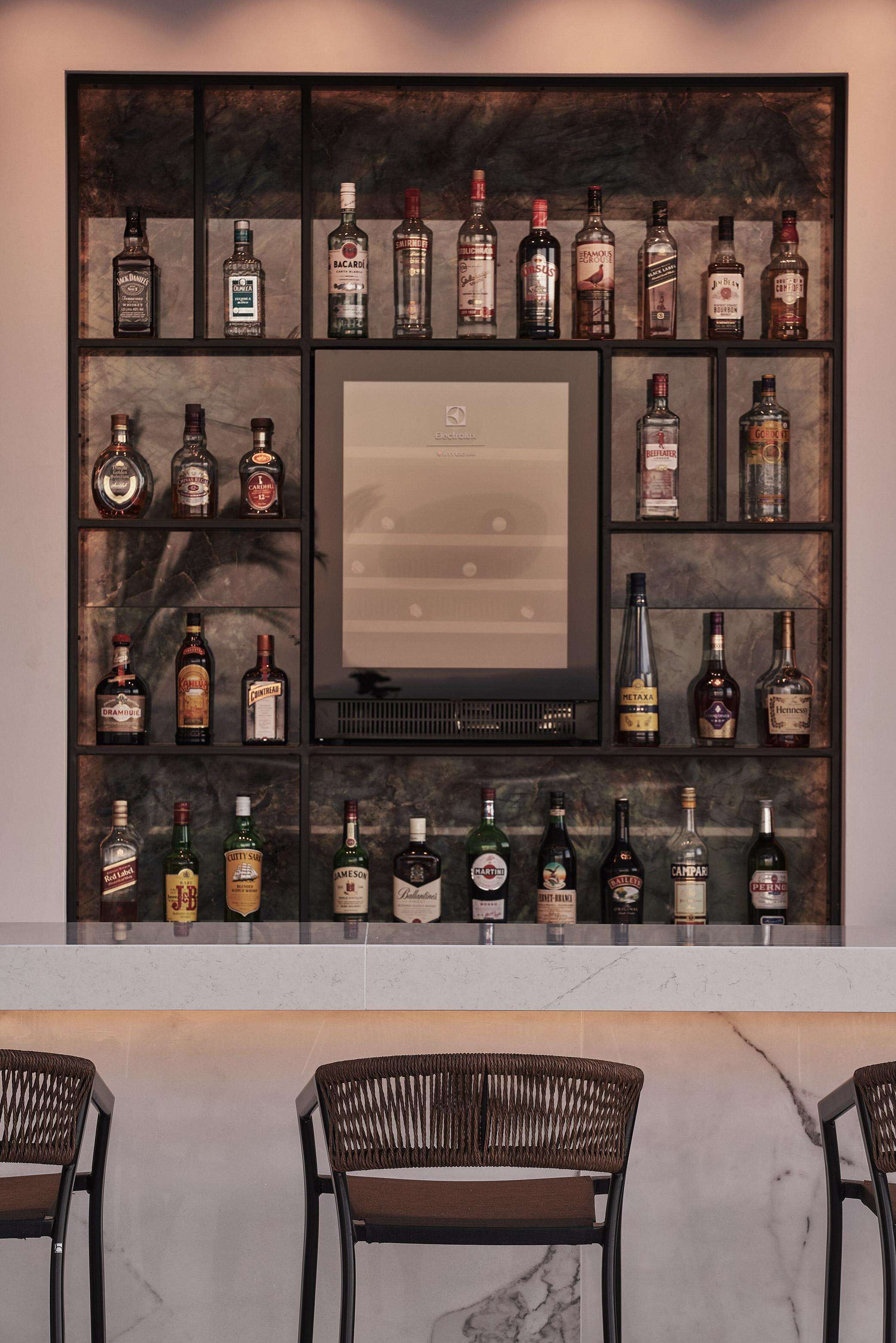 Una zona de bar con varias estanterías mostrando diferentes botellas de licor, una obra de arte enmarcada en el centro y tres sillas de mimbre.