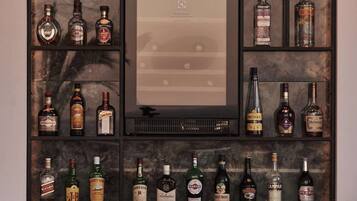 Una zona de bar con varias estanterías mostrando diferentes botellas de licor, una obra de arte enmarcada en el centro y tres sillas de mimbre.