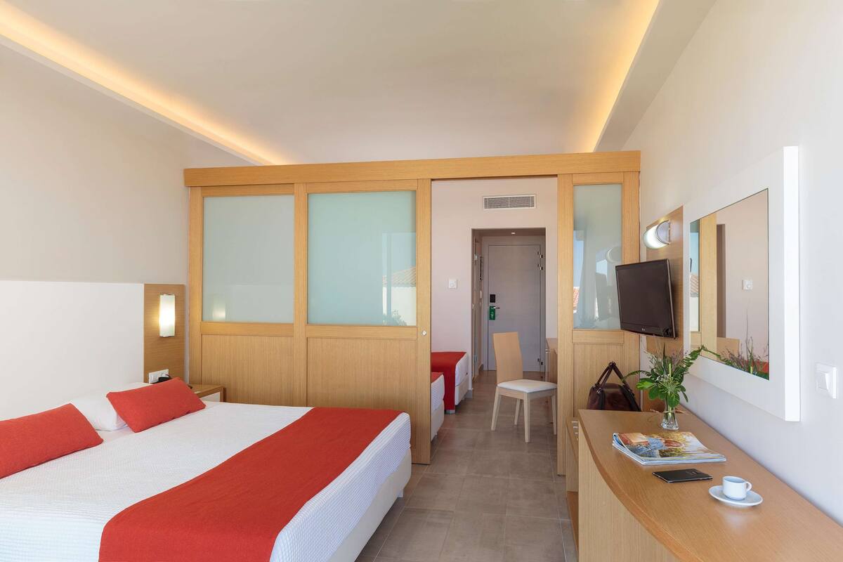 A hotel room with a large bed, a desk with a chair, a TV, and a small table with a cup and a book.