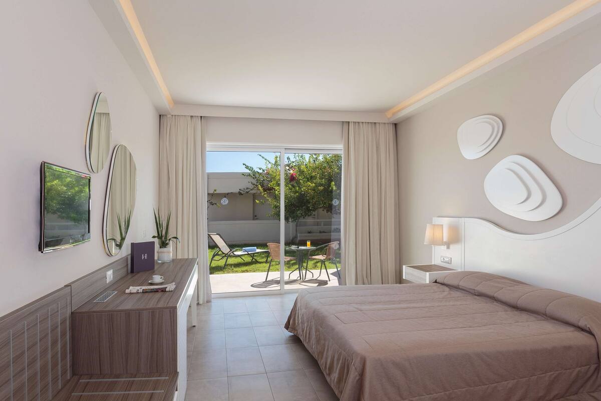 A modern hotel room with a large bed, a desk with a chair, a TV, and a sliding glass door to a balcony with outdoor seating.