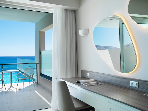 Une chambre d’hôtel moderne avec un balcon donnant sur la mer, un grand miroir et une coiffeuse en bois.