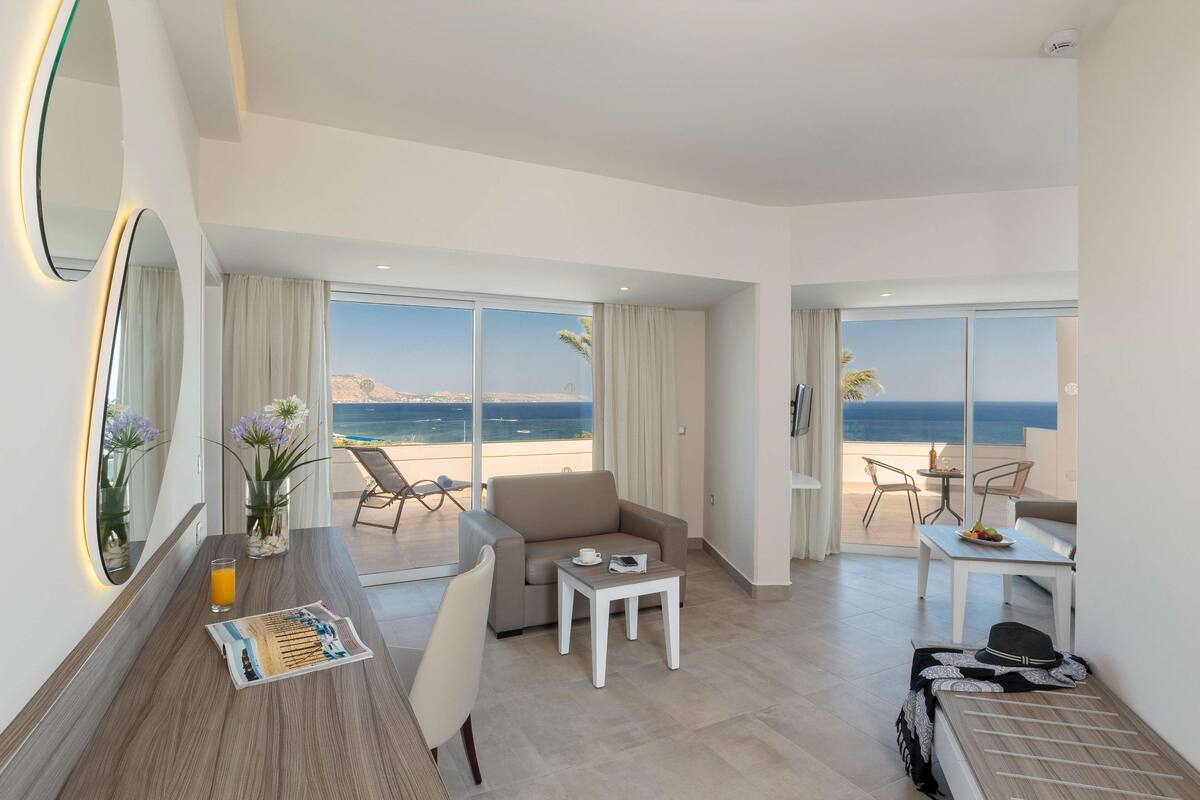 A modern hotel room with a large balcony, a sofa, a dining table, and a view of the beach.