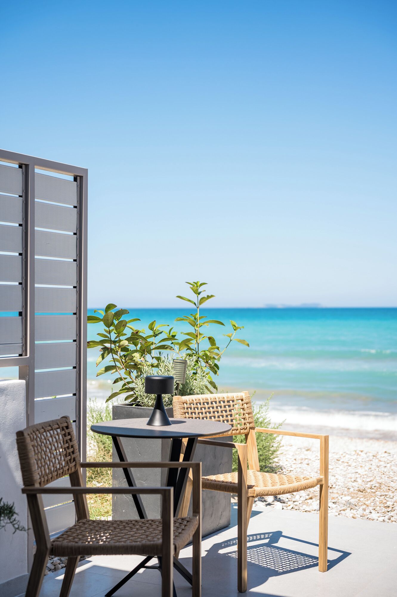 A patio with a table and chairs, overlooking a beach with clear blue water.