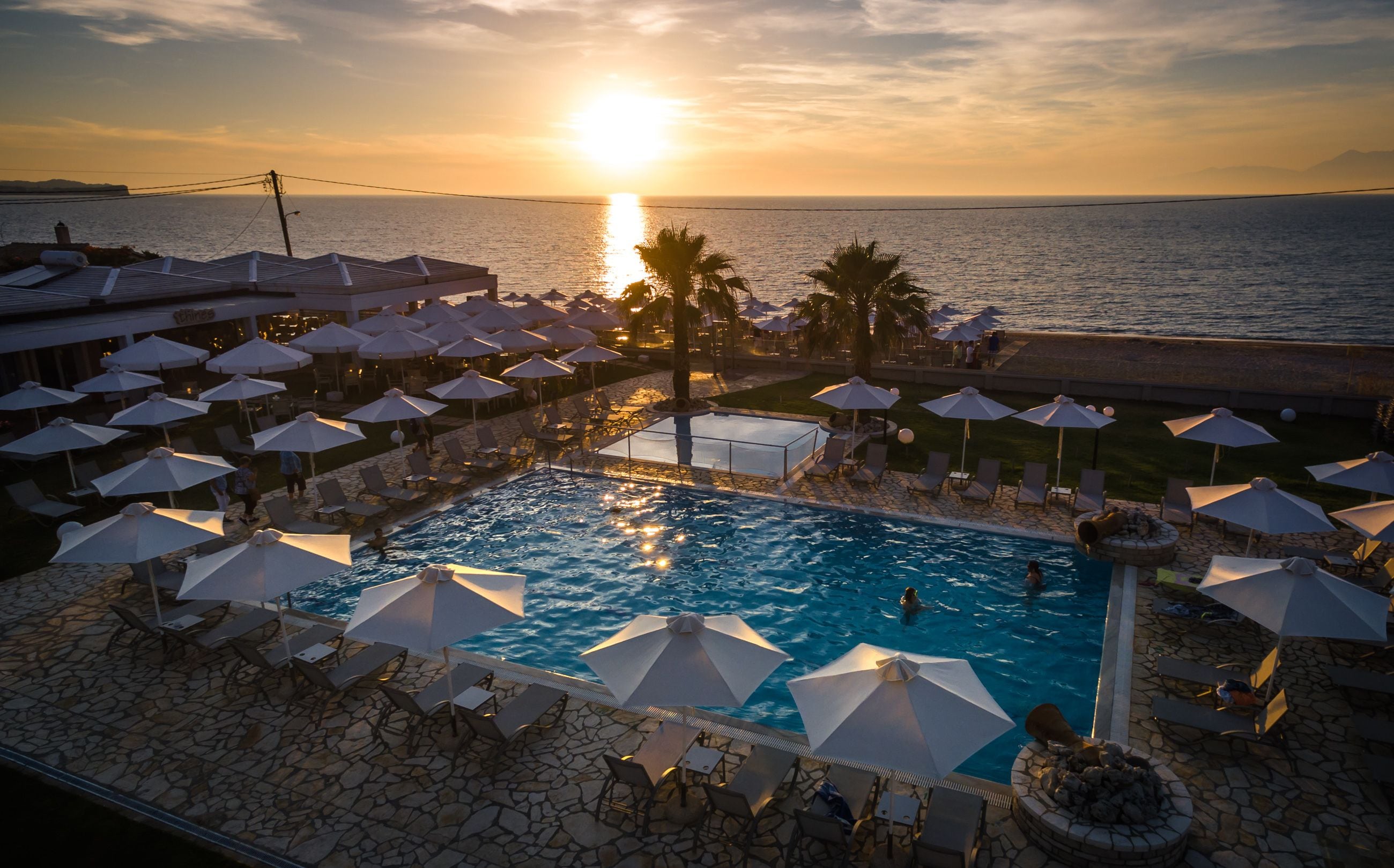 A poolside area with umbrellas and lounge chairs, overlooking a body of water at sunset.