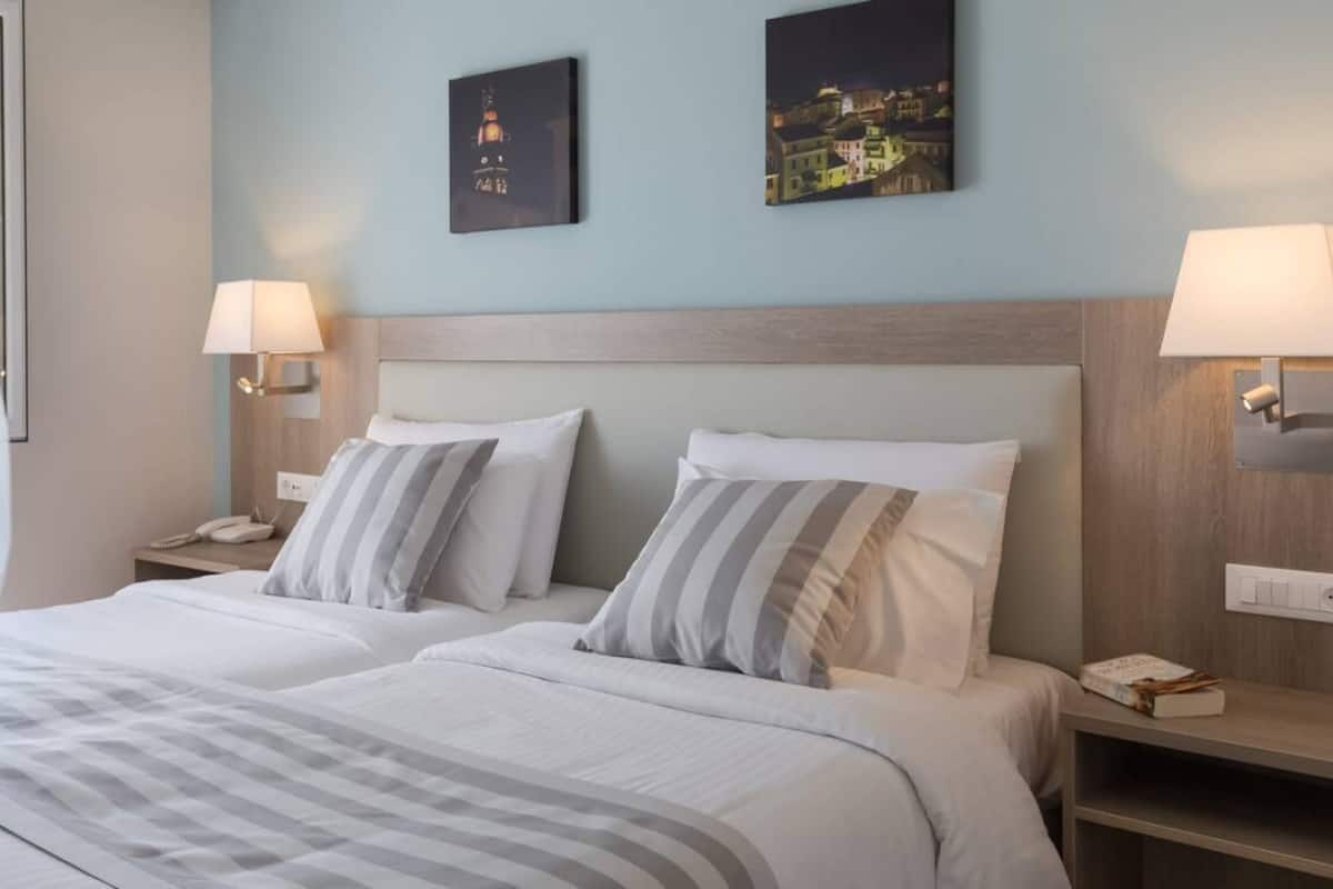 A hotel room with two beds, a nightstand with a lamp, a wall-mounted lamp, and two framed pictures.