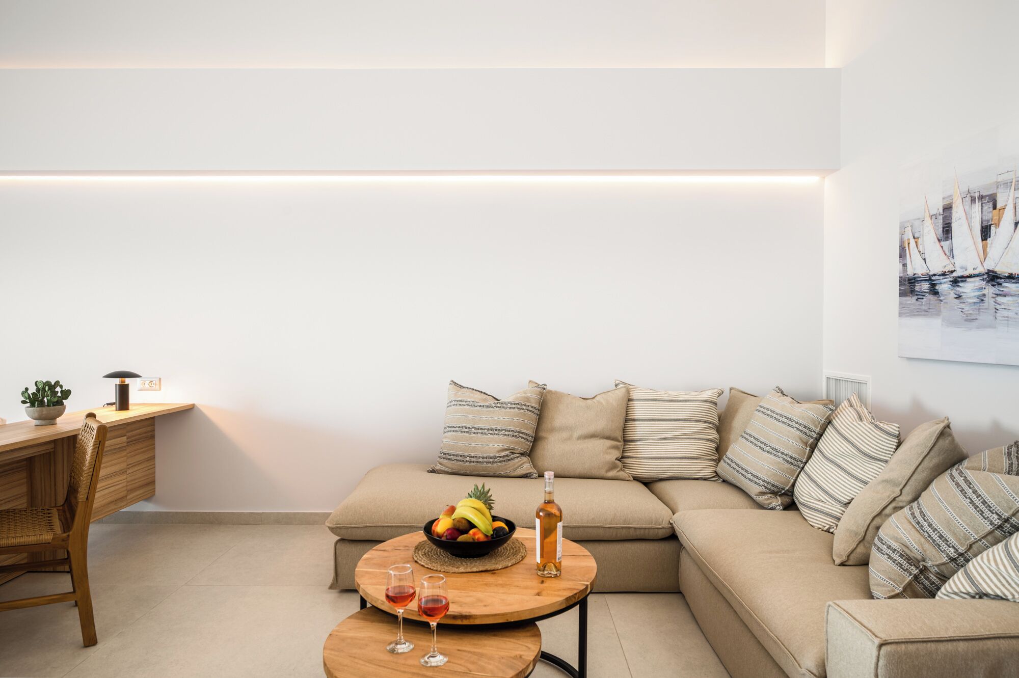 A modern living room with a wooden coffee table, a beige sofa with striped cushions, a fruit bowl, and two wine glasses.