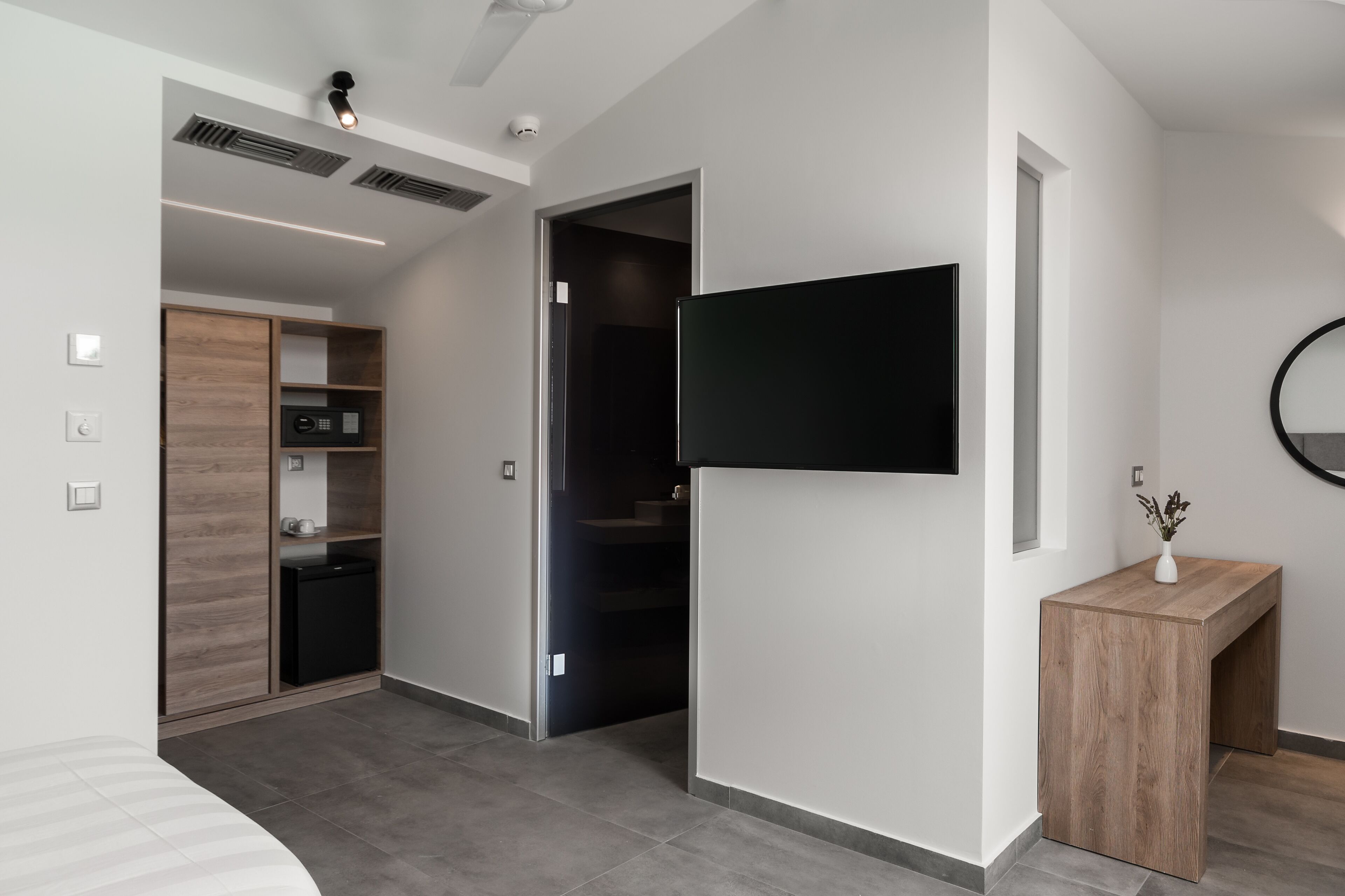 A modern hotel room with a flat-screen TV mounted on the wall, a wooden bedside table with a vase, and a built-in wardrobe with a small refrigerator.