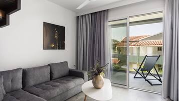 Una sala de estar moderna con un sofá gris, una mesa de centro blanca y una planta. Hay un ventanal corredero que da a un balcón con una silla y una mesa.